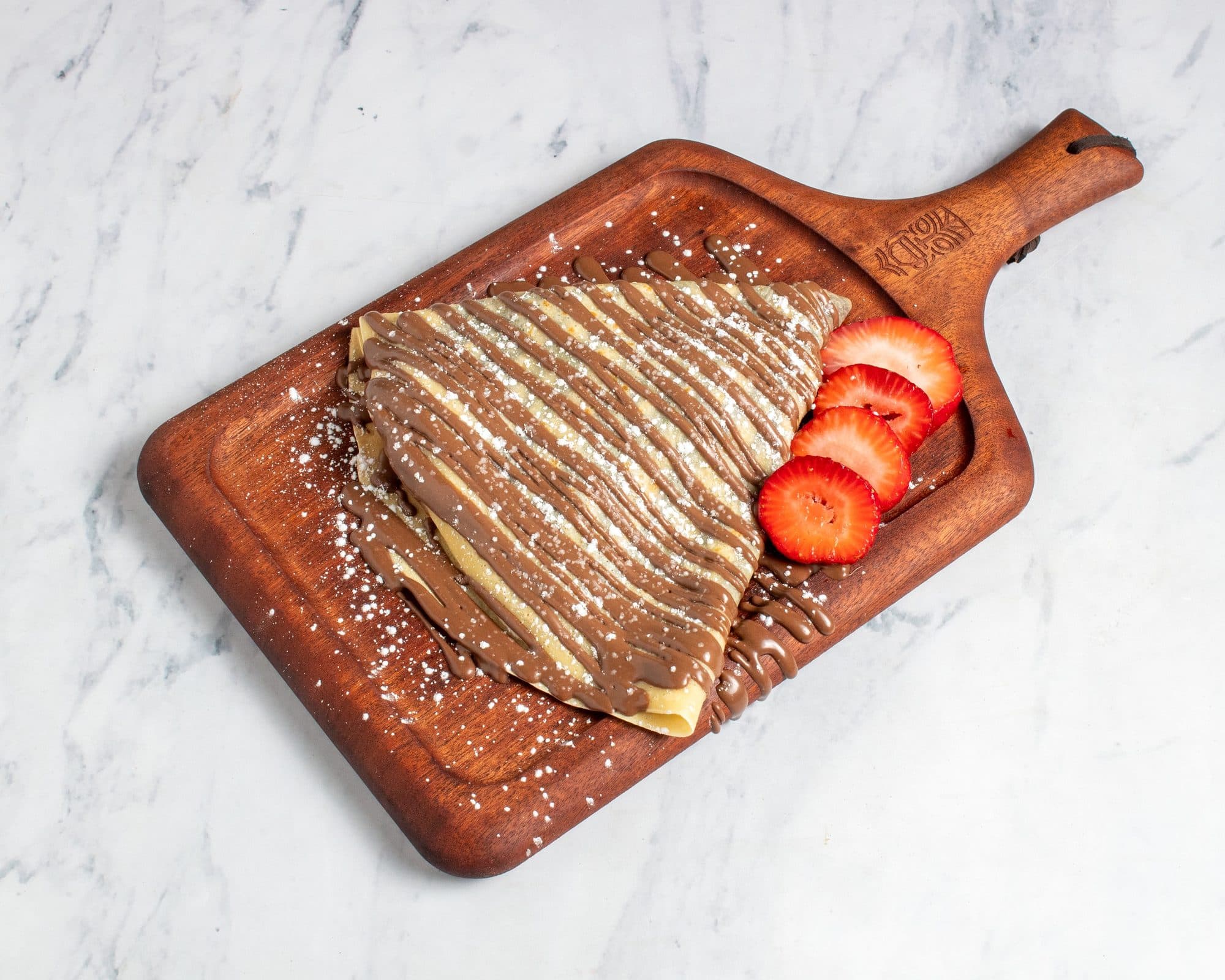 A folded crepe under chocolate drizzle and powdered sugar, with fresh strawberries